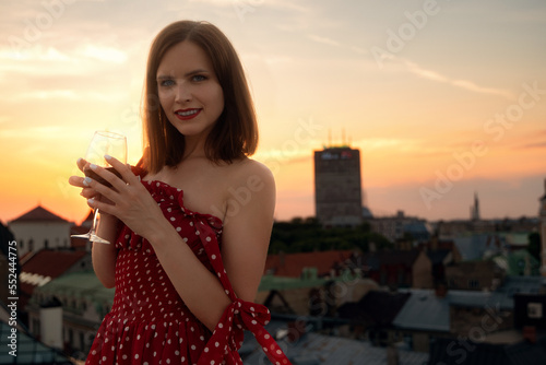 Young beautiful female model in the red dress enjoy the glass of wine on the roof of Old city of Riga, Latvia