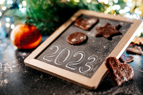 New Year sign 2023. Baking Christmas gingerbread cookies on a dark wooden table. Christmas lights in background. Winter holidays. Top view. Shallow depth of field. 