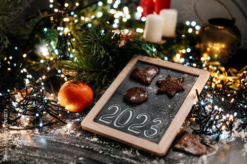 New Year sign 2023. Baking Christmas gingerbread cookies on a dark wooden table. Christmas lights in background. Winter holidays. Top view. Shallow depth of field. 