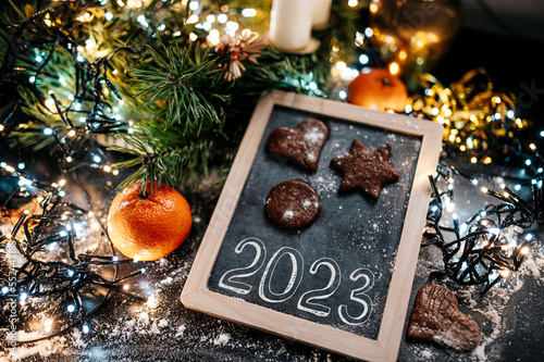 New Year sign 2023. Baking Christmas gingerbread cookies on a dark wooden table. Christmas lights in background. Winter holidays. Top view. Shallow depth of field. 