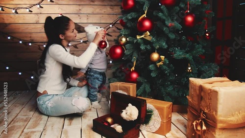 Mom and kid are decorating Christmas tree. Pretty multiracial Mother and toddler child at home decorate Christmas tree, put toys on branches. Christmas lights are shining in background. Slow motion photo