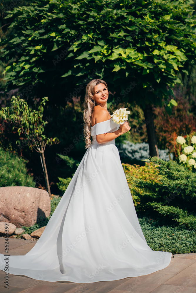 a blonde bride with bouquet Calla Lily in the park in a white long dress.