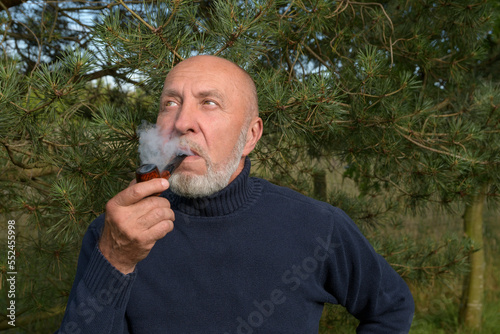 An aged man in a coniferous forest watches nature. A man with a smoking pipe enjoys the singing of birds. Lifestyle