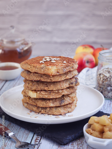 A plate of healthy rolled oat and apple pancakes with a cup of cooked diced apple pieces. Teapot set and its fruits in background with bottle of oat grains