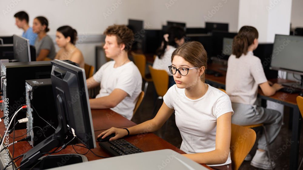 Positive female teenager in glasses learning computer science while she is using a PC in the computer classroom