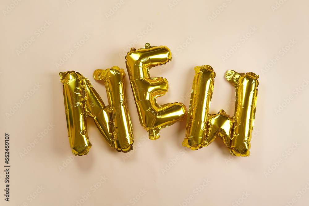 Huge golden foil letter balloons spelling new on beige colored seamless background, soft light shadow Huge golden foil letter balloons spelling new on beige colored seamless background, soft light shadow