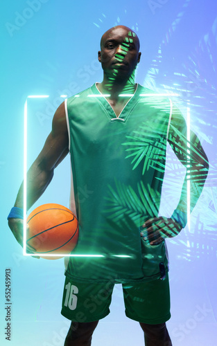Portrait of african american basketball player with ball standing by illuminated square and plants