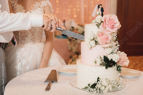 Newlyweds cut a three-tiered wedding cake decorated with rose flowers and a figurine of the bride and groom
