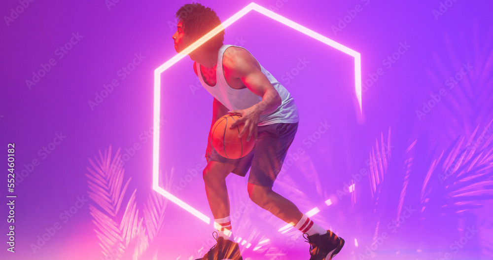 Biracial basketball player dribbling ball by illuminated hexagon and plants on violet background