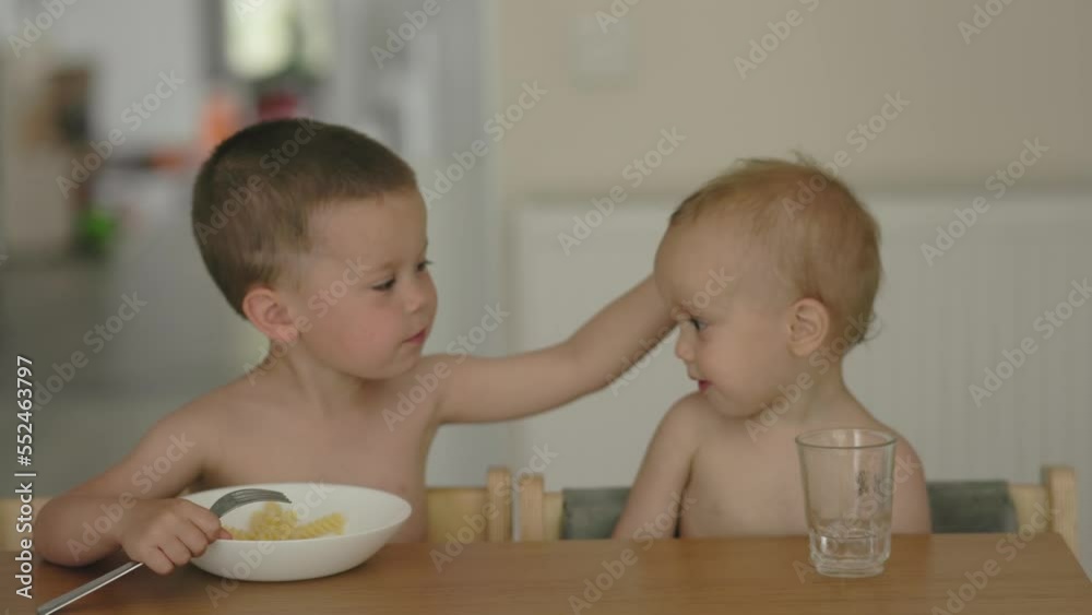 elder brother helping baby boy eat, petting child on head with hand. siblings eating together. loving kid caring his toddler baby brother patting his head. little friends in kitchen having meal elder brother helping baby boy eat, petting child on head with hand. siblings eating together. loving kid caring his toddler baby brother patting his head. little friends in kitchen having meal