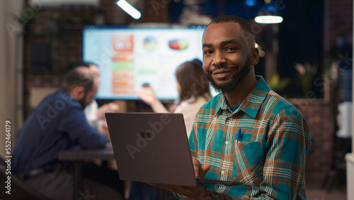 African american employee working on laptop, looking at camera, smiling. Young man holding computer medium shot portrait, business meeting statistics presentation in background. Handheld shot.