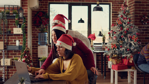 Multiethnic team of women doing teamwork in festive space, wearing santa hats and working on report. Brainstorming new startup ideas together during christmas holiday season with decorations.