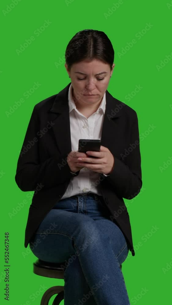 Vertical video: Front view close up of businesswoman browsing internet on mobile phone, acting casual on chair in studio. Company worker in suit using smartphone app to text messages, full body Vertical video: Front view close up of businesswoman browsing internet on mobile phone, acting casual on chair in studio. Company worker in suit using smartphone app to text messages, full body
