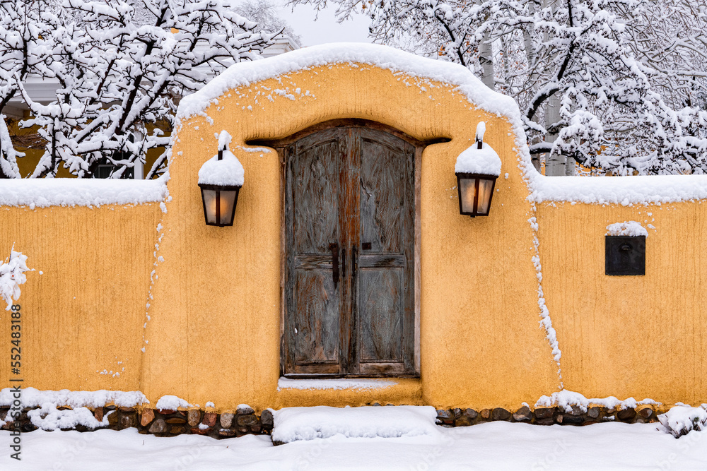 Naklejka premium Snowy scene in Santa Fe of adobe wall, glowing lights and rustic door