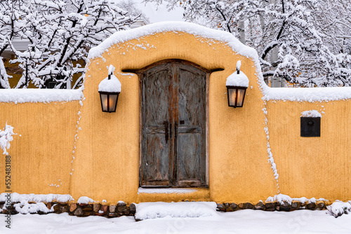 Snowy scene in Santa Fe of adobe wall, glowing lights and rustic door