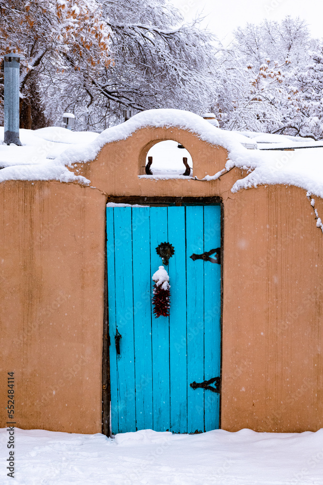 Obraz premium Winter scene of snow-covered adobe wall with a turquoise colored door and red chile ristra in Santa Fe, New Mexico