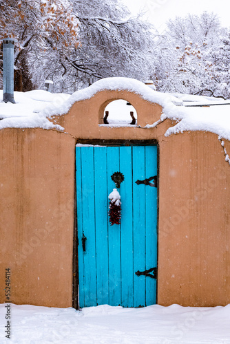 Winter scene of snow-covered adobe wall with a turquoise colored door and red chile ristra in Santa Fe, New Mexico
