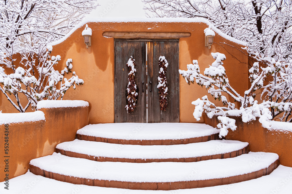 Fototapeta premium Winter scene of snow-covered adobe wall with rustic wood doors and chile ristras in Santa Fe, New Mexico