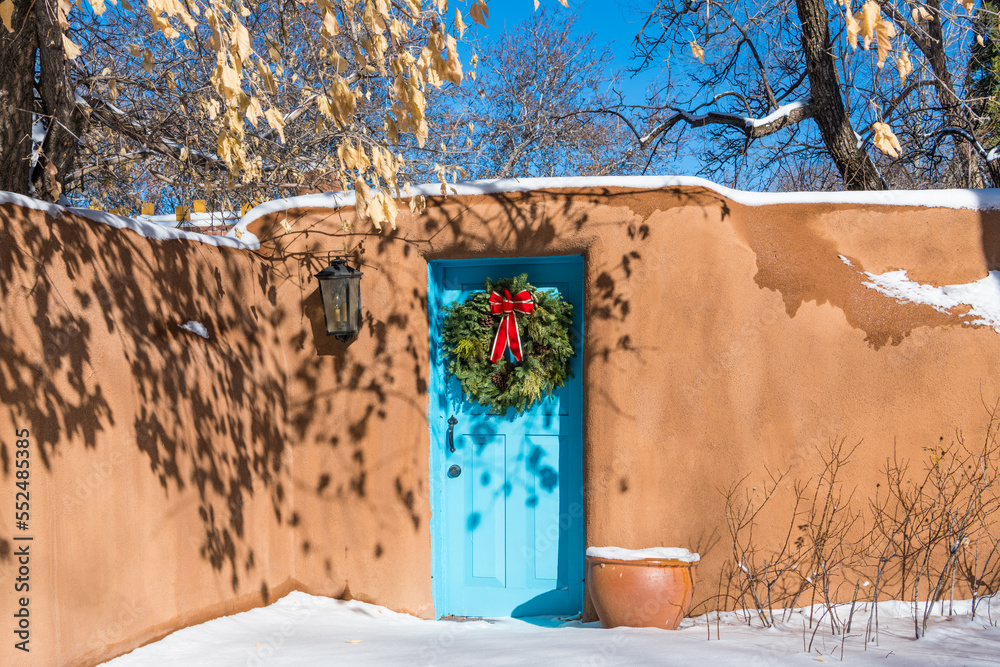 Fototapeta premium Snowy winter scene of Christmas wreath on turquoise colored door in adobe wall in Santa Fe, New Mexico