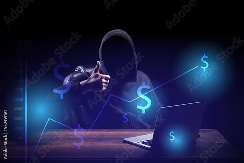 An online investor, dressed in a black hood, sits in a dark room with a laptop on the table.