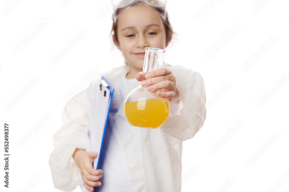 Selective focus on laboratory flask with yellow liquid chemical, in hands of smart elementary school student in lab coat, holding clipboard, showing chemical reaction taking place in flask to camera