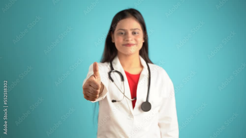 Portrait of a young female doctor gesturing thumbs up, Cheerful Asian Indian woman doctor in apron and stethoscope isolated over blue studio background Portrait of a young female doctor gesturing thumbs up, Cheerful Asian Indian woman doctor in apron and stethoscope isolated over blue studio background