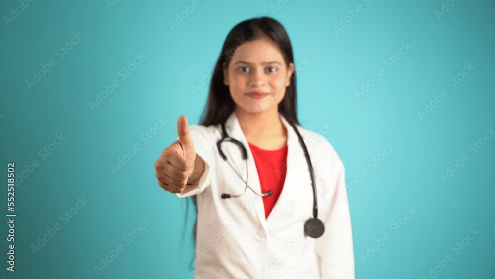 Portrait of a young female doctor gesturing thumbs up, Cheerful Asian Indian woman doctor in apron and stethoscope isolated over blue studio background