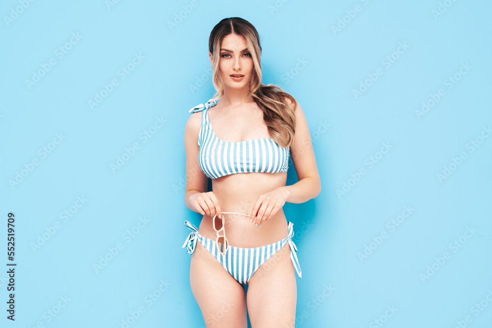 Fototapeta premium Young smiling blond model in summer swimwear colourful bathing suit. Sexy carefree woman having fun and going crazy. Female posing near blue wall in studio. Cheerful and happy. Perfect body