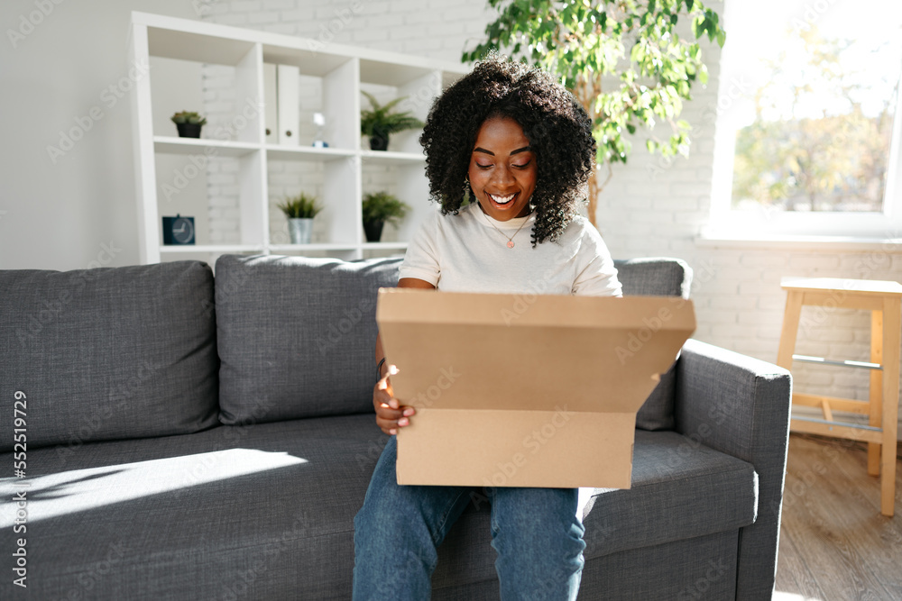 Young african woman sit on couch at home unpacking parcel cardboard box with online purchase Young african woman sit on couch at home unpacking parcel cardboard box with online purchase