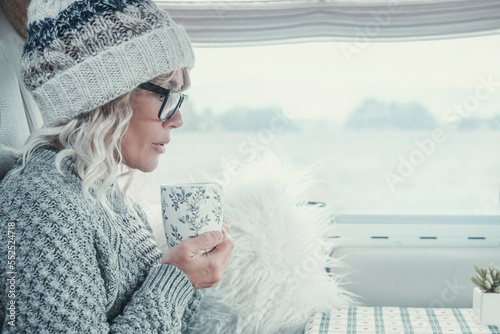 Side view of attractive lady with white hair and clothes drinking from a cup inside her camper van during winter season. Bright image of serene woman. Portrait of female people in inside leisure photo