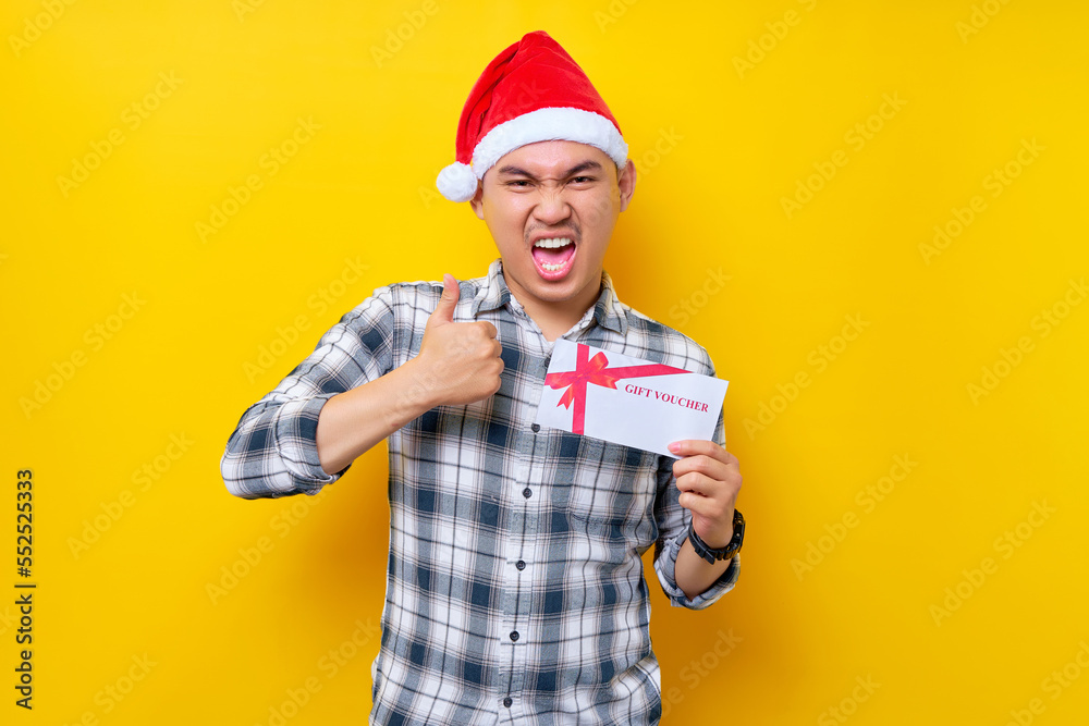 Smiling cheerful young Asian man 20s in a Santa Claus hat holding a gift certificate coupon voucher card for the store, showing thumb up looking camera isolated on yellow studio background Smiling cheerful young Asian man 20s in a Santa Claus hat holding a gift certificate coupon voucher card for the store, showing thumb up looking camera isolated on yellow studio background