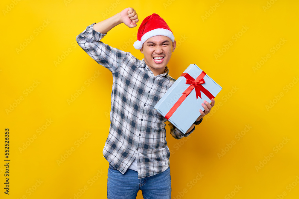 Excited young Asian man with a Santa Claus hat Showing a gift box and celebrating luck isolated on yellow studio background. Merry Christmas Concept Excited young Asian man with a Santa Claus hat Showing a gift box and celebrating luck isolated on yellow studio background. Merry Christmas Concept