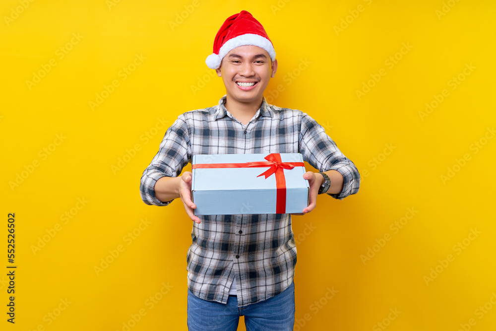Excited young Asian man wearing plaid shirt with a Santa Claus hat giving a gift box isolated on yellow studio background. Merry Christmas Concept Excited young Asian man wearing plaid shirt with a Santa Claus hat giving a gift box isolated on yellow studio background. Merry Christmas Concept