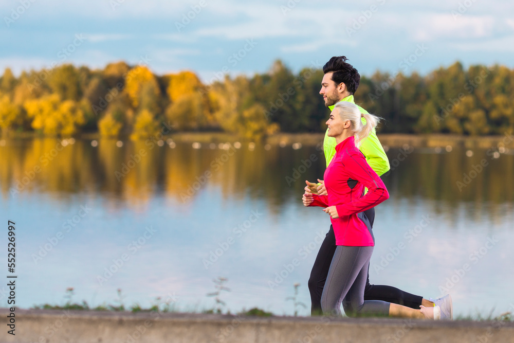 Jogging Concepts. Running Couple While Training Outdoors on Road At River For Durable Marathon Pretty Young Female and Extremely Fit Caucasian Handsome Man