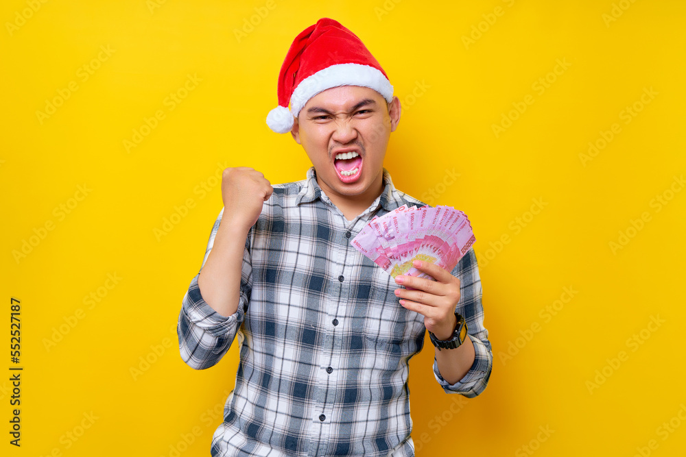 Ecited young Asian man 20s wearing a Christmas hat holding cash money rupiah and Raising Fist, Celebrating victory isolated on yellow background