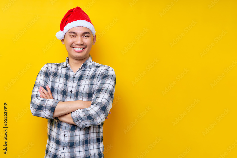 Smiling young Asian man wearing a plaid shirt in a Christmas hat standing hold hands crossed and folded looks camera on yellow background. celebration Christmas holiday and New Year concept