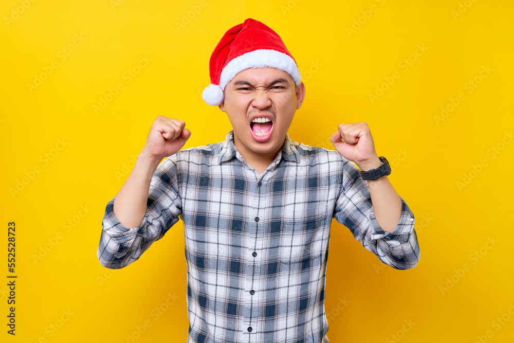 Smiling young Asian man wearing a plaid shirt in a Christmas hat posing doing winner gesture clenching fists on yellow background. celebration Christmas holiday and New Year concept Smiling young Asian man wearing a plaid shirt in a Christmas hat posing doing winner gesture clenching fists on yellow background. celebration Christmas holiday and New Year concept