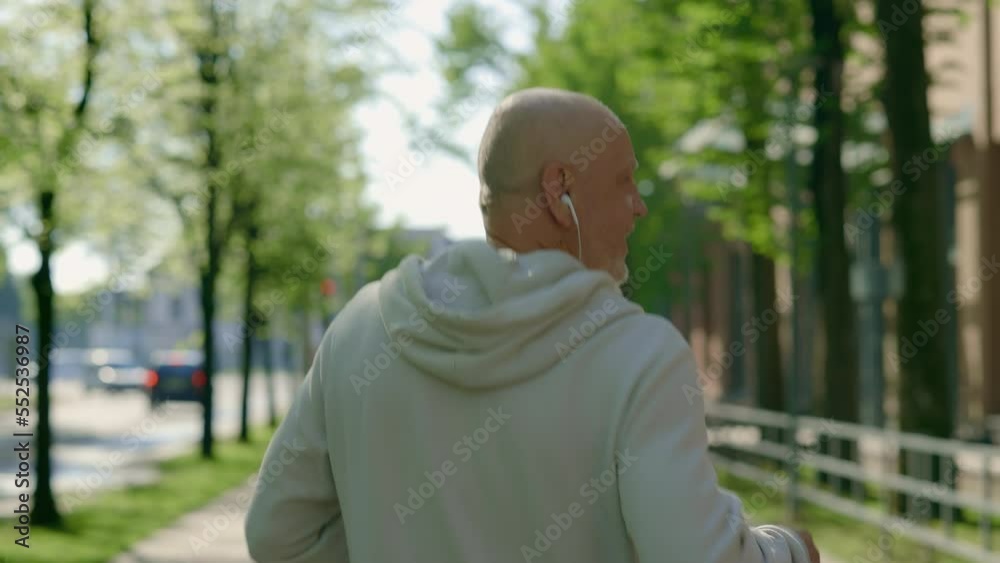Back view of the mature man running through the street wearing the headphones. Sport and healthy lifestyle concept. Aging Youthfully Back view of the mature man running through the street wearing the headphones. Sport and healthy lifestyle concept. Aging Youthfully