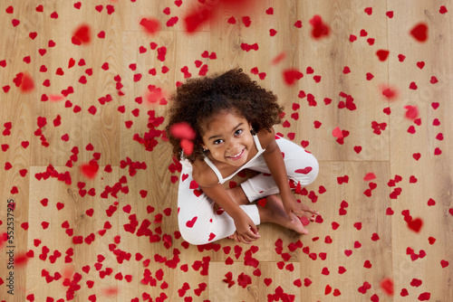 Top view of smiling young girl sitting on floor surrounded by heart shaped confetti