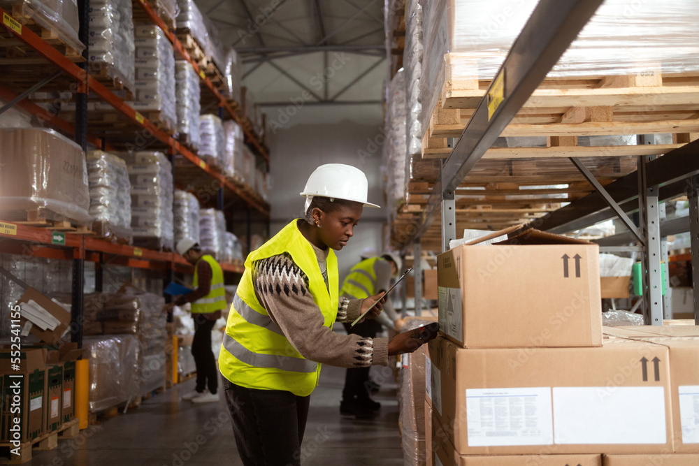 An African-American woman in protective workwear is working in a distribution warehouse taking inventory and moving packages.. She is scanning the boxes with a barcode reader. An African-American woman in protective workwear is working in a distribution warehouse taking inventory and moving packages.. She is scanning the boxes with a barcode reader.
