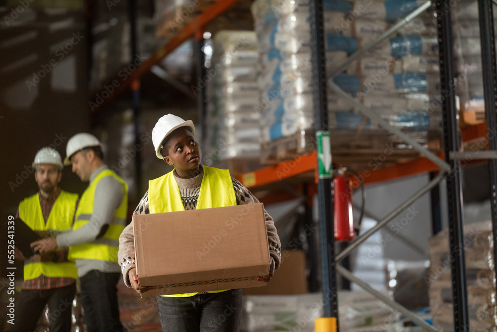 A female warehouse worker wearing protective clothing is carrying a box in a large warehouse.