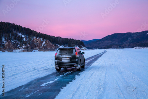 Winter travel. The car drives along an icy road along a frozen river bed against the backdrop of mountains and a sunset sky.