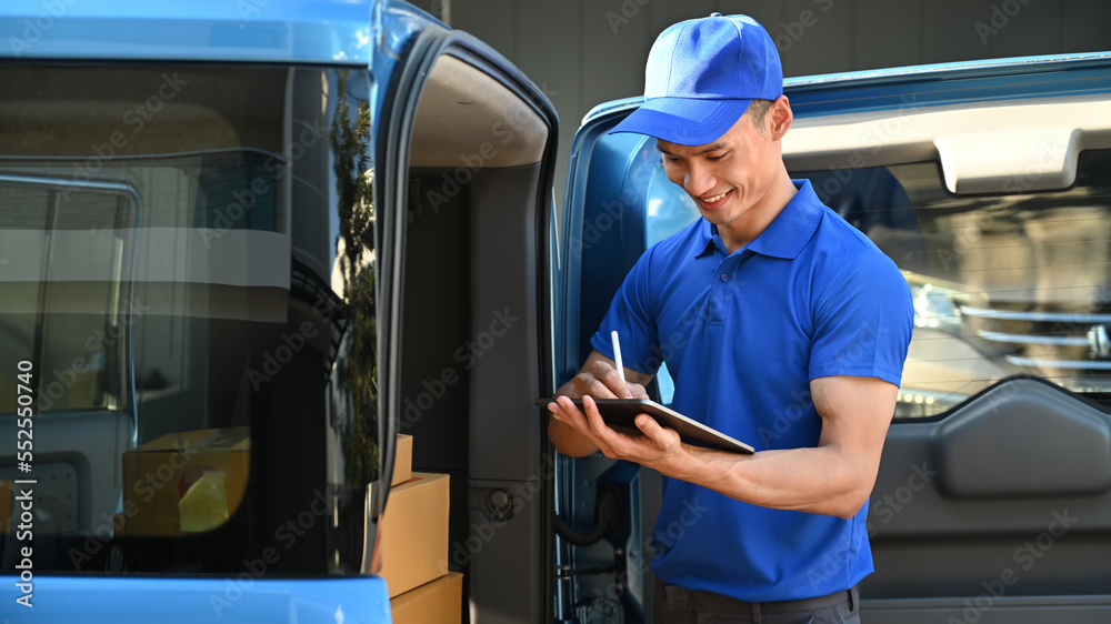 Smiling asian male courier in blue uniform using digital tablet next to open delivery van. Shipping and delivery service concept Smiling asian male courier in blue uniform using digital tablet next to open delivery van. Shipping and delivery service concept