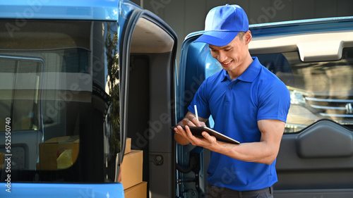 Smiling asian male courier in blue uniform using digital tablet next to open delivery van. Shipping and delivery service concept
