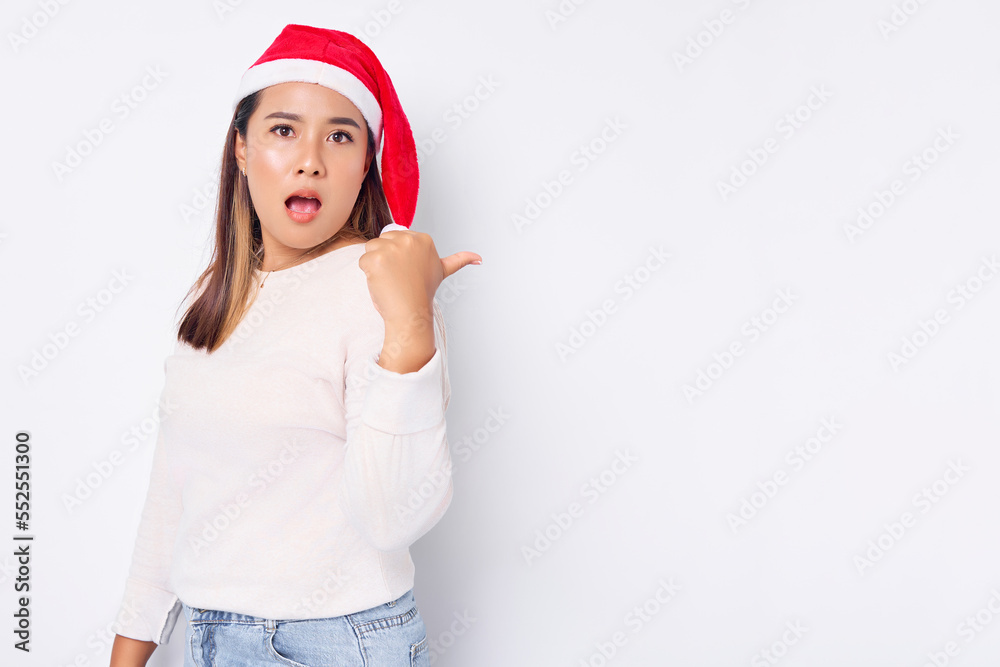 Surprised young Asian woman in a Santa Claus hat pointing thumb at copy space isolated over white background. celebration Christmas holiday and New Year concept Surprised young Asian woman in a Santa Claus hat pointing thumb at copy space isolated over white background. celebration Christmas holiday and New Year concept
