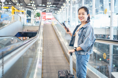 Photography Asian woman with mobile phone in hand and luggage standing on escalator traveler at international airport, young girl tourist lifestyle on holidays