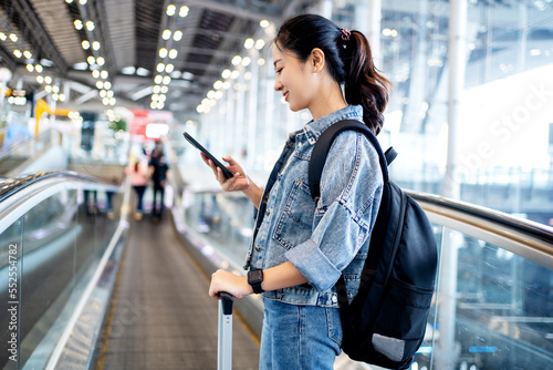 Asian woman tourist in casual clothes using a smartphone on escalator at airport terminal. New normal travel at airport