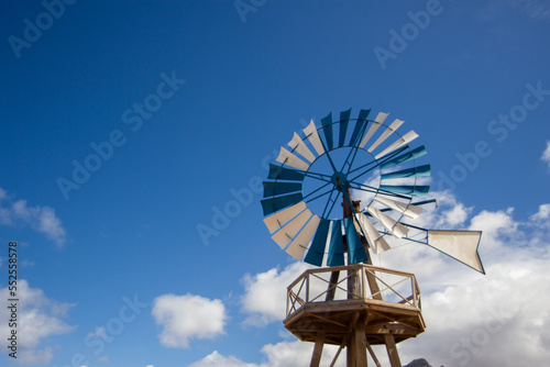 Desertic landscape. Windmill with blue and white blades. Mountains in the background. Yellow sand dunes with desert plants. Blue sky with white fluffy clouds. Famara Beach, Lanzarote, Canary Islands, 