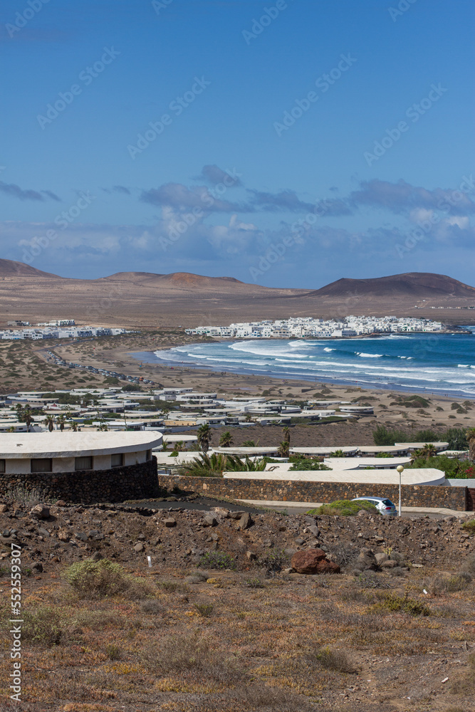 View of Caleta de Famara from the bungalows. Houses. Desertic landscape. Beach with waves, village and mountains in the background. Clear sky. Famara Beach, Lanzarote, Canary Islands, Spain.