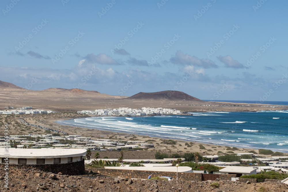 View of Caleta de Famara from the bungalows. Houses. Desertic landscape. Beach with waves, village and mountains in the background. Clear sky. Famara Beach, Lanzarote, Canary Islands, Spain. View of Caleta de Famara from the bungalows. Houses. Desertic landscape. Beach with waves, village and mountains in the background. Clear sky. Famara Beach, Lanzarote, Canary Islands, Spain.